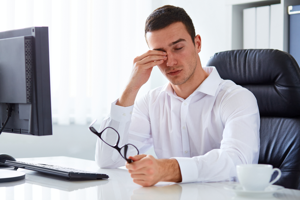 Guy sitting at desk wiping eyes with hand. 